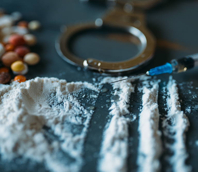 A pile of pills, a syringe, and other drugs on a table next to handcuffs.