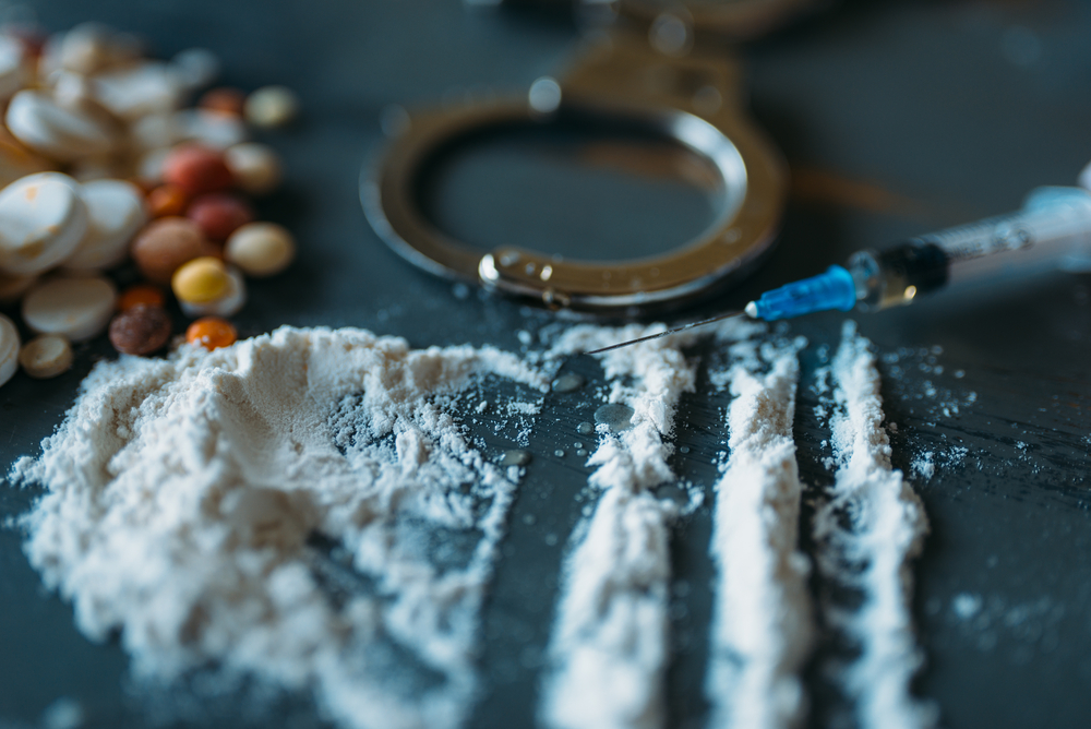 A pile of pills, a syringe, and other drugs on a table next to handcuffs.
