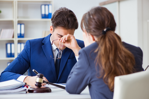 A man in a suit with his glasses off, looking frustrated, and a woman sitting across from him.