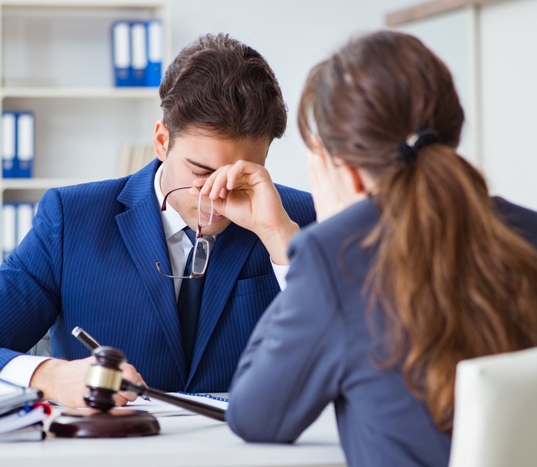 A man in a suit with his glasses off, looking frustrated, and a woman sitting across from him.