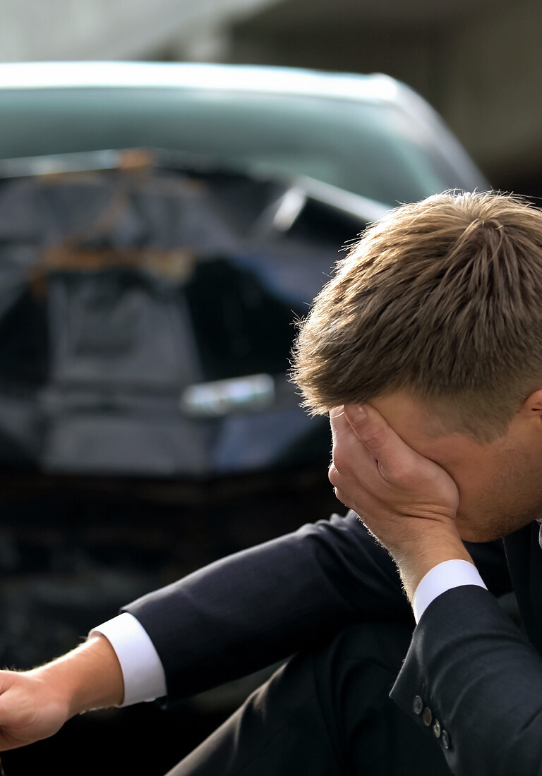 Drunk upset male driver holding bottle sitting street on crashed car background